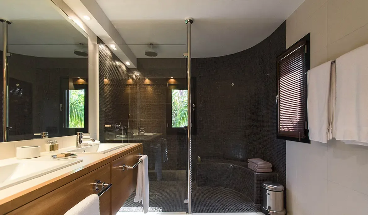 A curved mosaic shower with a built-in bench pairs with a warm wood double vanity under a massive mirror, creating a spa-like bathroom with lush garden views through a side window.