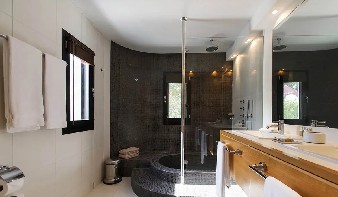 A sunken circular shower with dark mosaic tiles and a glass partition steals the spotlight, while a warm wood vanity with dual sinks and a frameless mirror keeps the rest bright and serene.