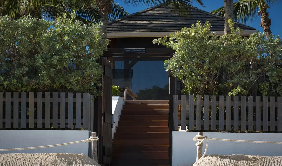 Wide wooden stairs lead through a private gate to the beach, framed by lush coastal greenery and a white picket fence atop a stone wall.