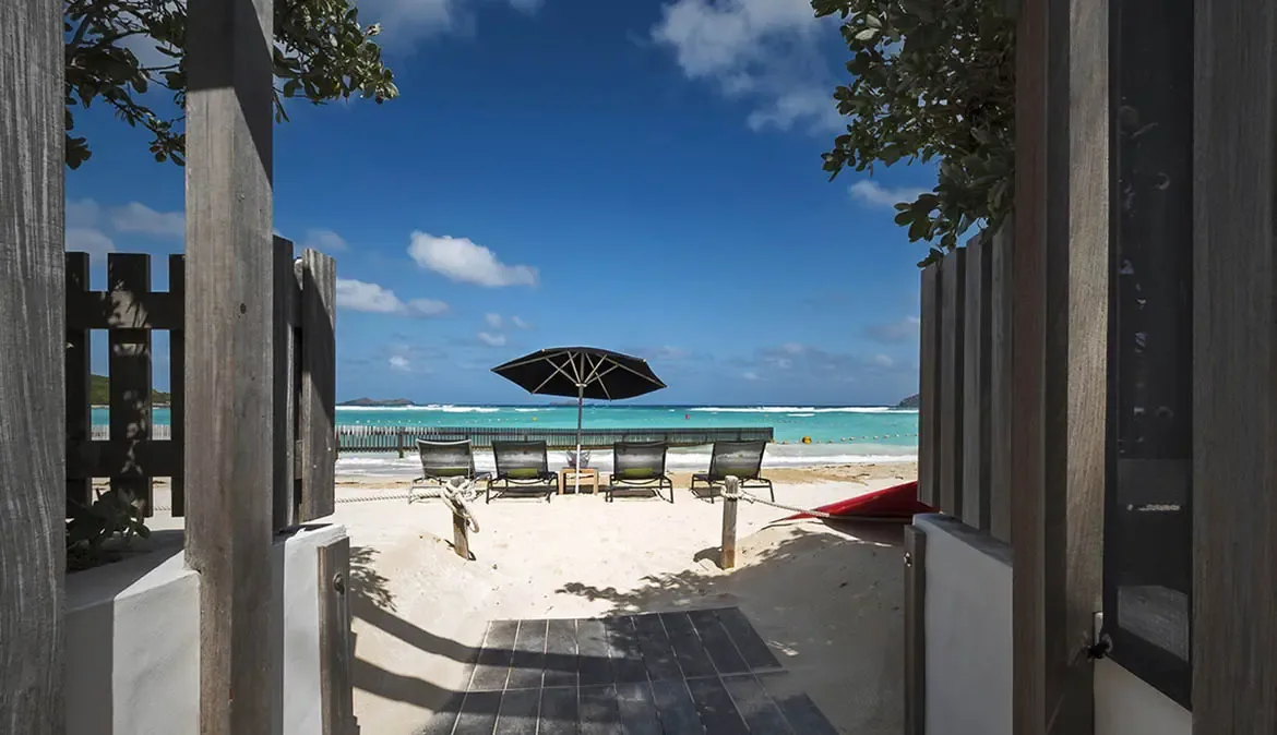 A black octagonal umbrella shades four loungers on white sand, with an ice bucket between chairs and turquoise Caribbean water just steps away.