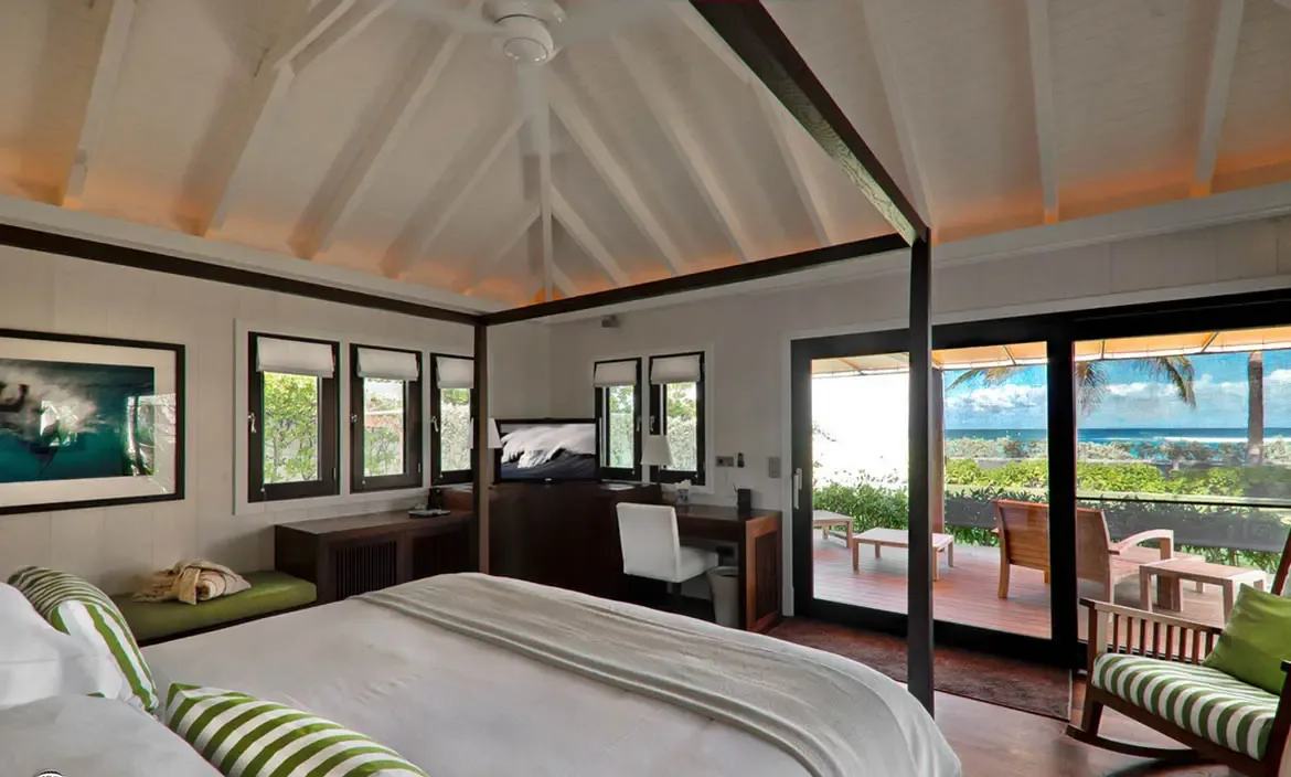 A dark wood canopy bed with green-striped pillows sits under vaulted white rafters, where open doors reveal a wooden deck with loungers and a turquoise ocean beyond.