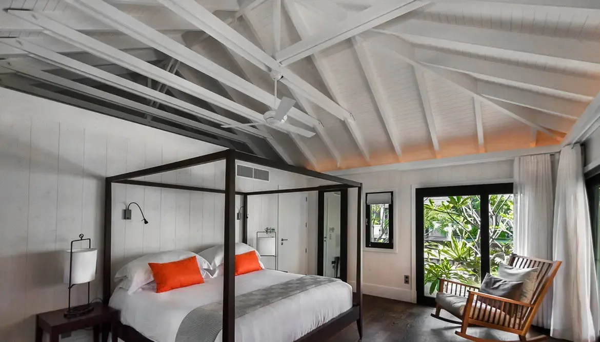 An orange pillow accents this canopy bedroom with dark vaulted rafters, where a wooden rocking chair by the glass doors invites quiet moments overlooking the tropical garden.