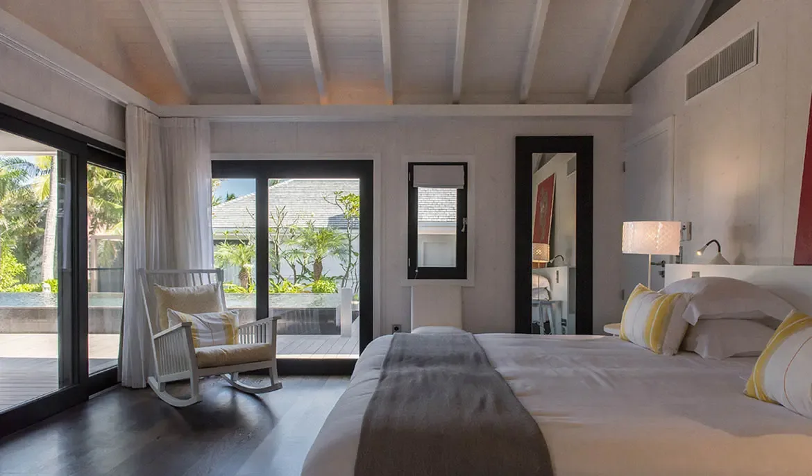 A dark wood canopy bed with yellow-striped pillows sits under vaulted white rafters, where a rocking chair and glass doors open to a pool deck and tropical greenery.
