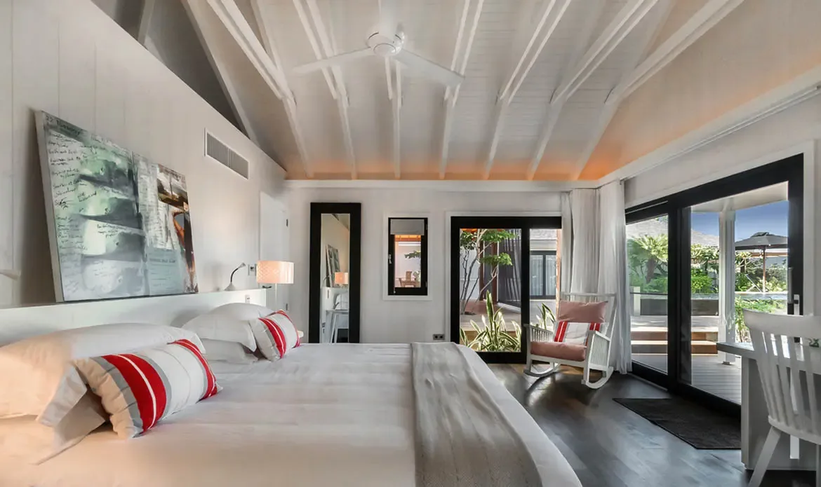Red and grey striped pillows add warmth to a white bed under vaulted rafters, where a rocking chair by glass doors opens to tropical gardens and a deck beyond.