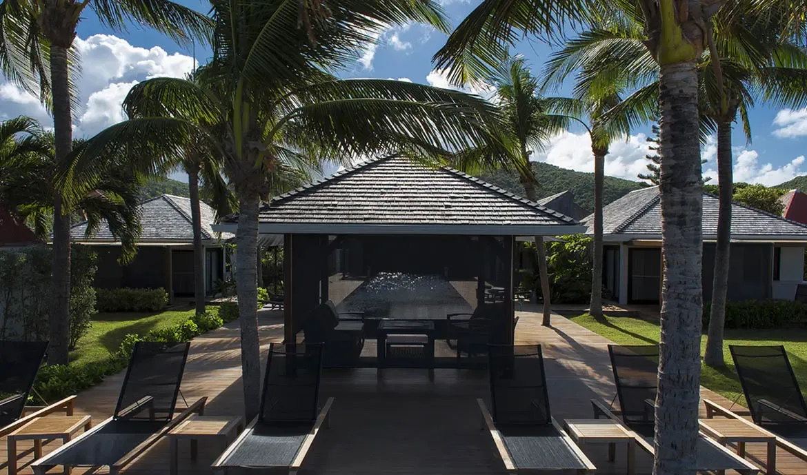 Sleek loungers and a coffee table sit on a wide wooden deck, where a shingled gazebo and palm trees frame a bright Caribbean day under a sky of fluffy white clouds.