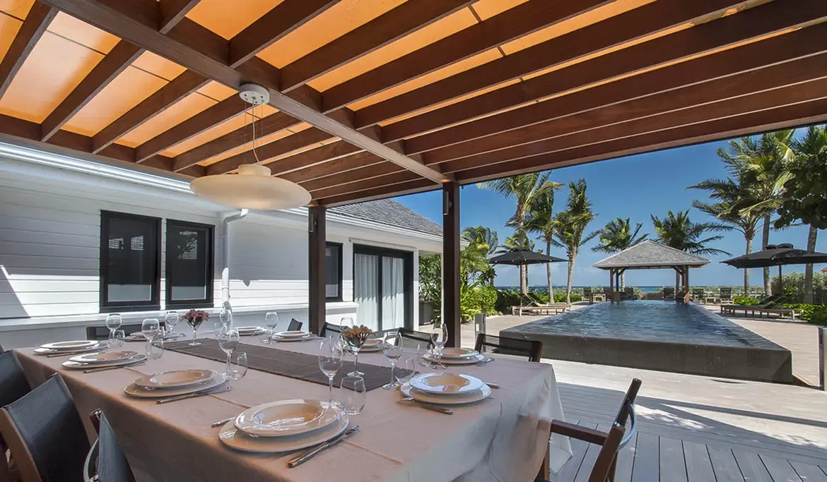 A formally set dining table with white linens and silverware sits under a warm-glow pergola, while an infinity pool and ocean stretch toward a gazebo on the far deck.