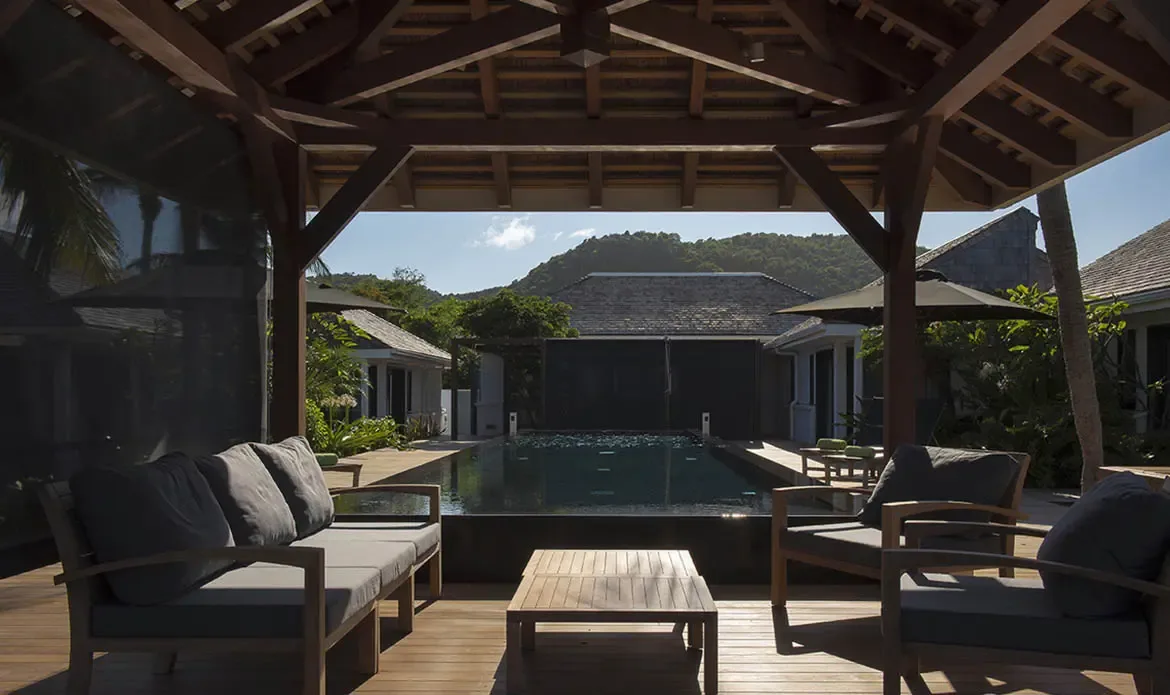 Dark cushions on a sleek sectional fill a shaded gazebo, where the open frame reveals a dark-tiled pool, tropical gardens, and a forested mountain under a bright blue sky.