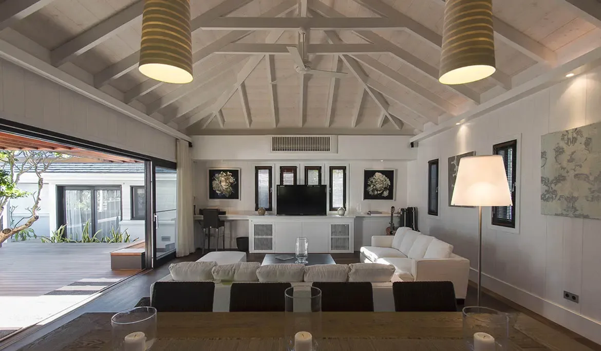 A plush L-shaped white sofa and dark coffee table sit under vaulted white rafters, where sliding glass doors open wide to a wooden deck and lush tropical greenery.