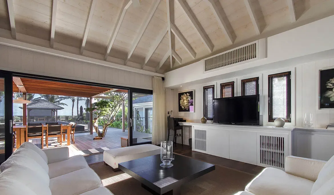 A white L-shaped sectional faces a built-in TV console in this vaulted living room, where open doors lead to an outdoor dining pergola and tropical gardens.