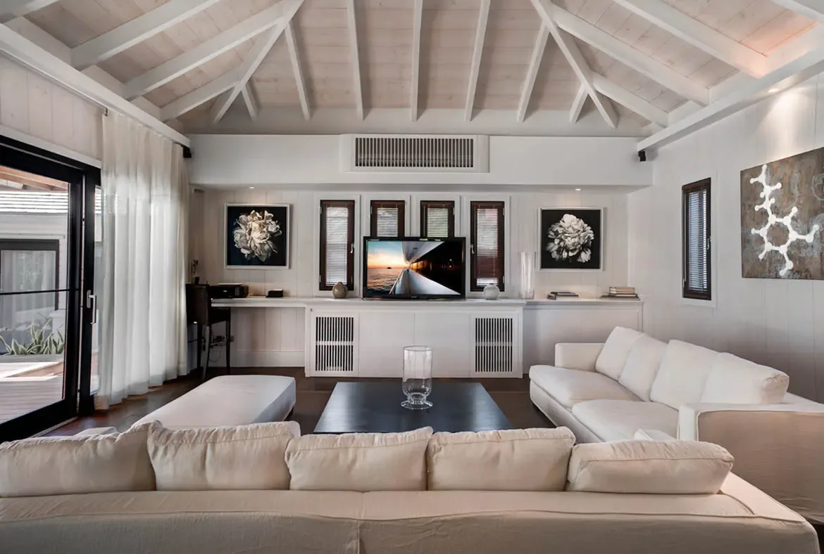 An all-white sectional anchors this vaulted living room with dark hardwood floors, where a built-in TV wall and glass doors open to the tropical deck beyond.
