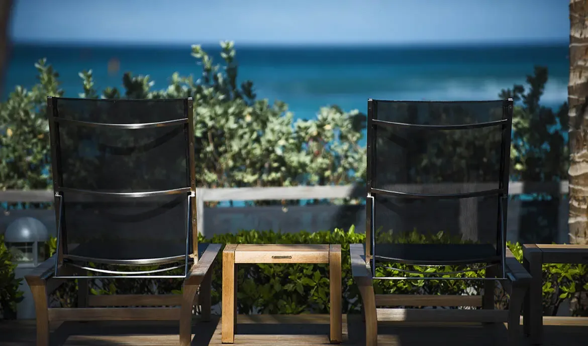 Two sleek lounge chairs face the ocean on a wooden deck, with lush tropical foliage creating a natural green screen between you and the deep blue Caribbean.