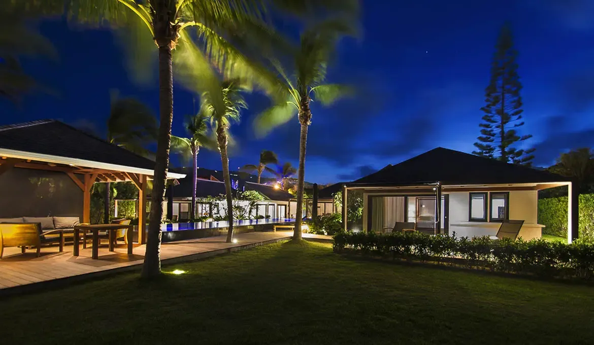 At dusk, uplit palms and glowing pavilion interiors illuminate a mirrored infinity pool, where warm amber and cool blue create a cinematic Caribbean evening.