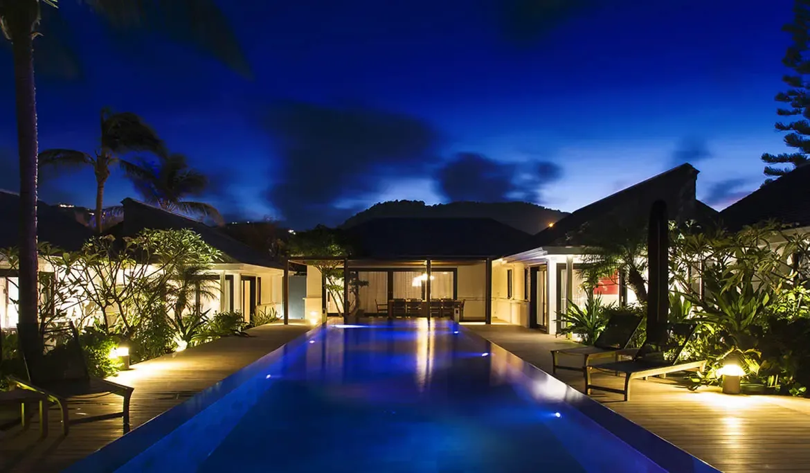 At dusk, warm interior lights and cool blue pool reflections create a cinematic scene, where pavilions with glass walls glow beside an infinity lap pool under an indigo sky.