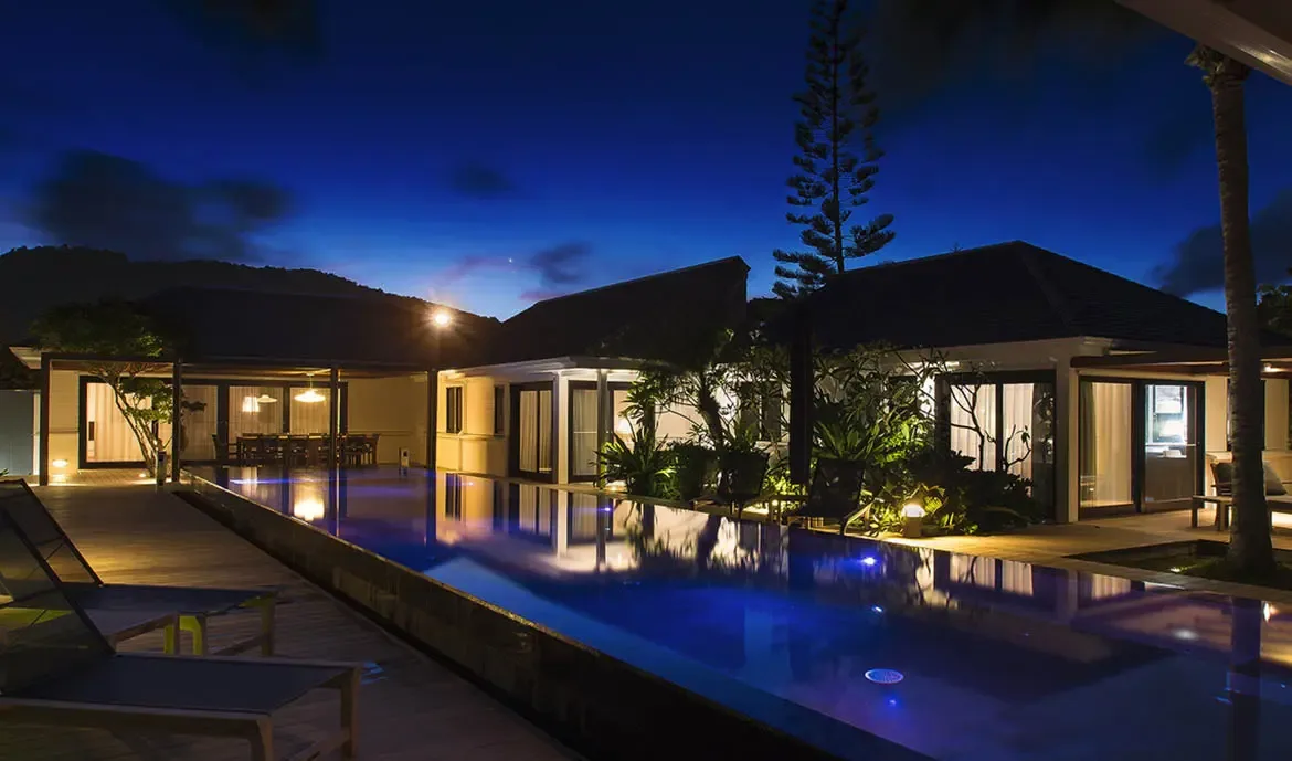 At twilight, an infinity pool mirrors the warm glow of pavilion interiors, while palm silhouettes and a dark evergreen rise against a deep indigo sky over forested mountains.