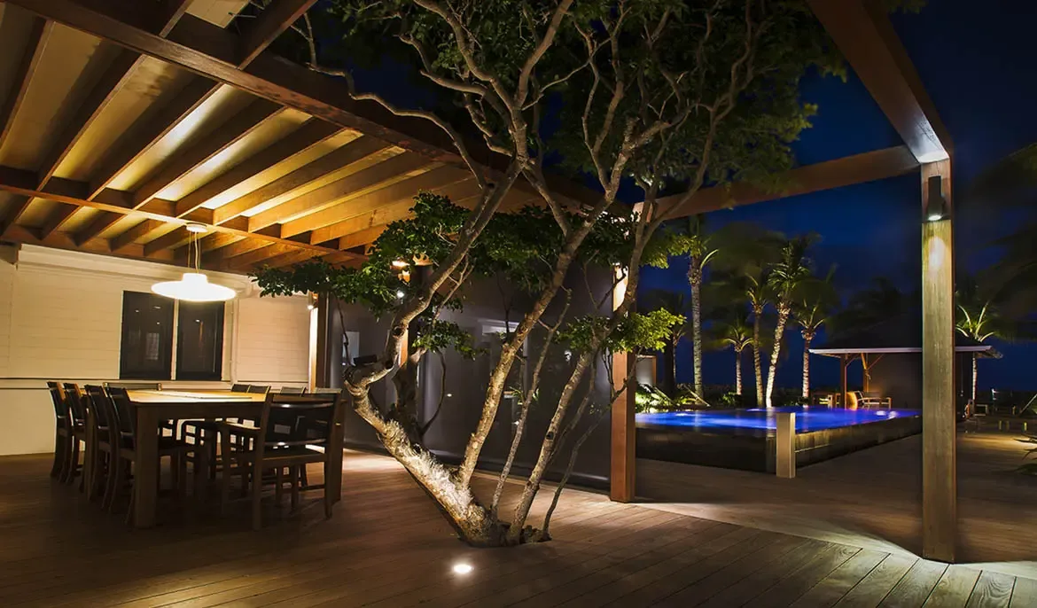 At night, warm uplighting transforms a tree growing through the deck into a sculptural centerpiece beside a glowing infinity pool and an illuminated outdoor dining pergola.
