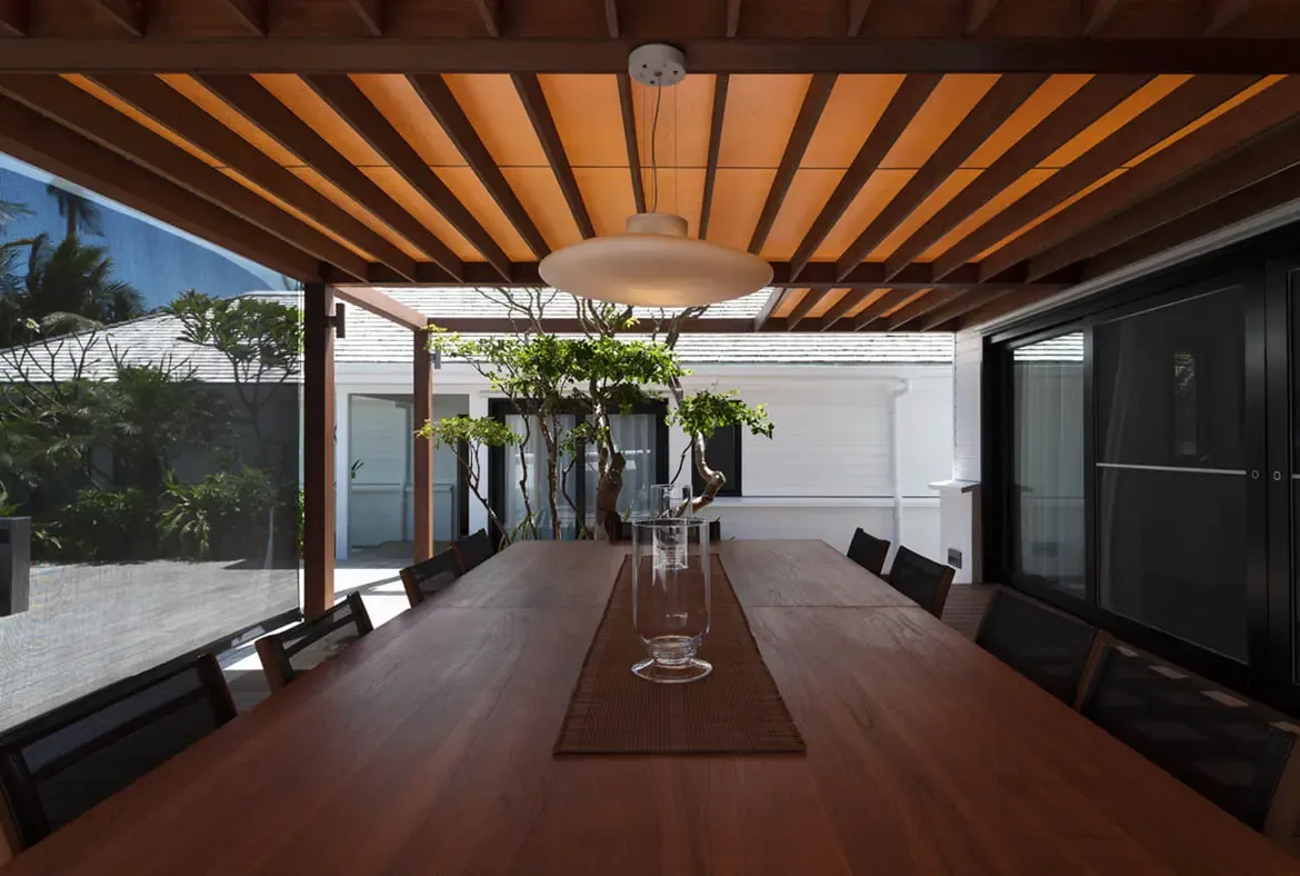 A long wooden dining table sits under a slatted pergola with warm amber panels and a sleek disc pendant light, opening to a courtyard of lush greenery and white-walled pavilions.