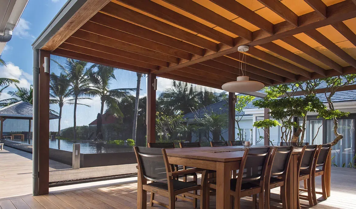 A modern dining pavilion with a white disc pendant light opens to an infinity pool and tropical gardens, where palm trees and manicured hedges frame a sun-drenched Caribbean retreat.