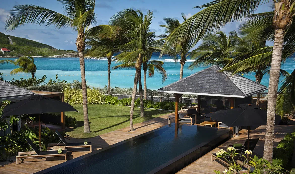 From above, a dark-tiled infinity pool stretches toward a turquoise bay, with green-towel loungers, a shingled gazebo, and palm trees creating a perfect beachfront compound.