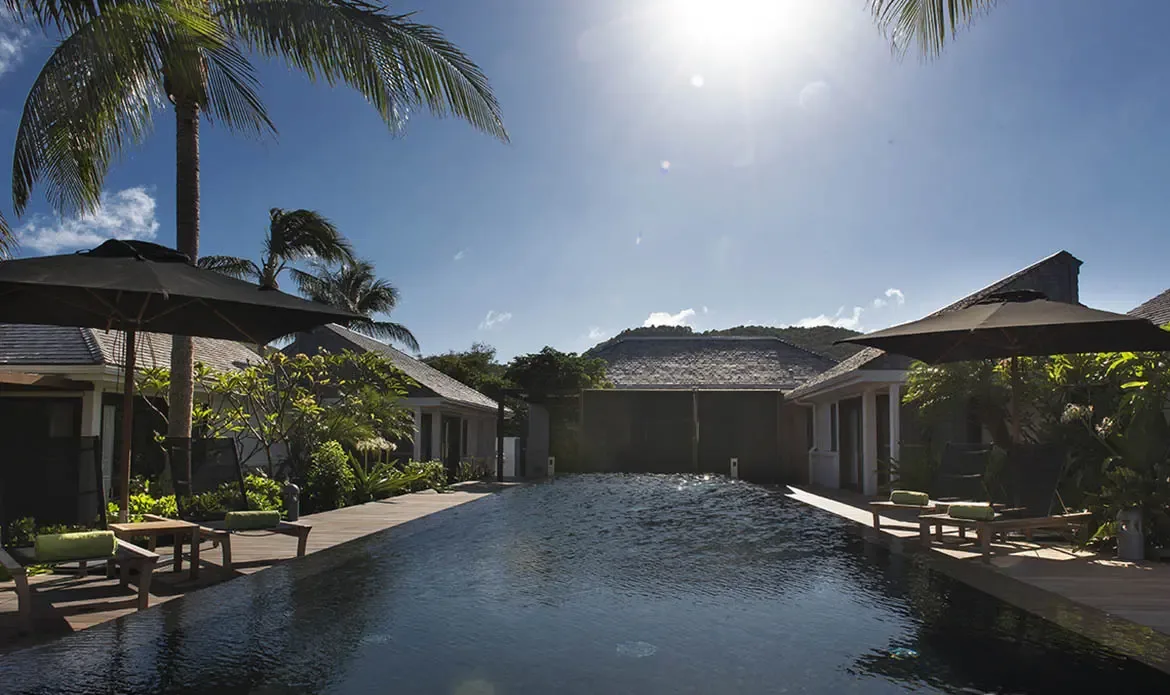 An aerial view captures a dark-tiled pool with green-towel loungers and black umbrellas, surrounded by white pavilions and lush palms against a forested green hill.