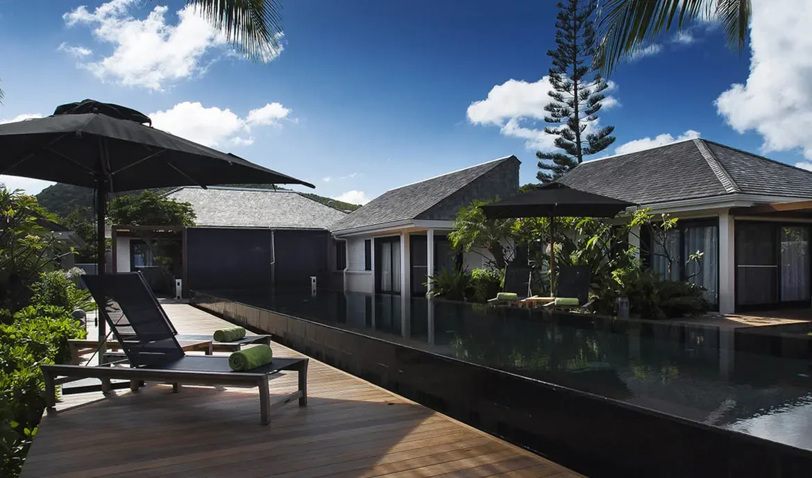 Green-towel loungers line a dark-tiled infinity pool under black umbrellas, where white pavilions with grey-shingled roofs create a private tropical compound under a brilliant sky.