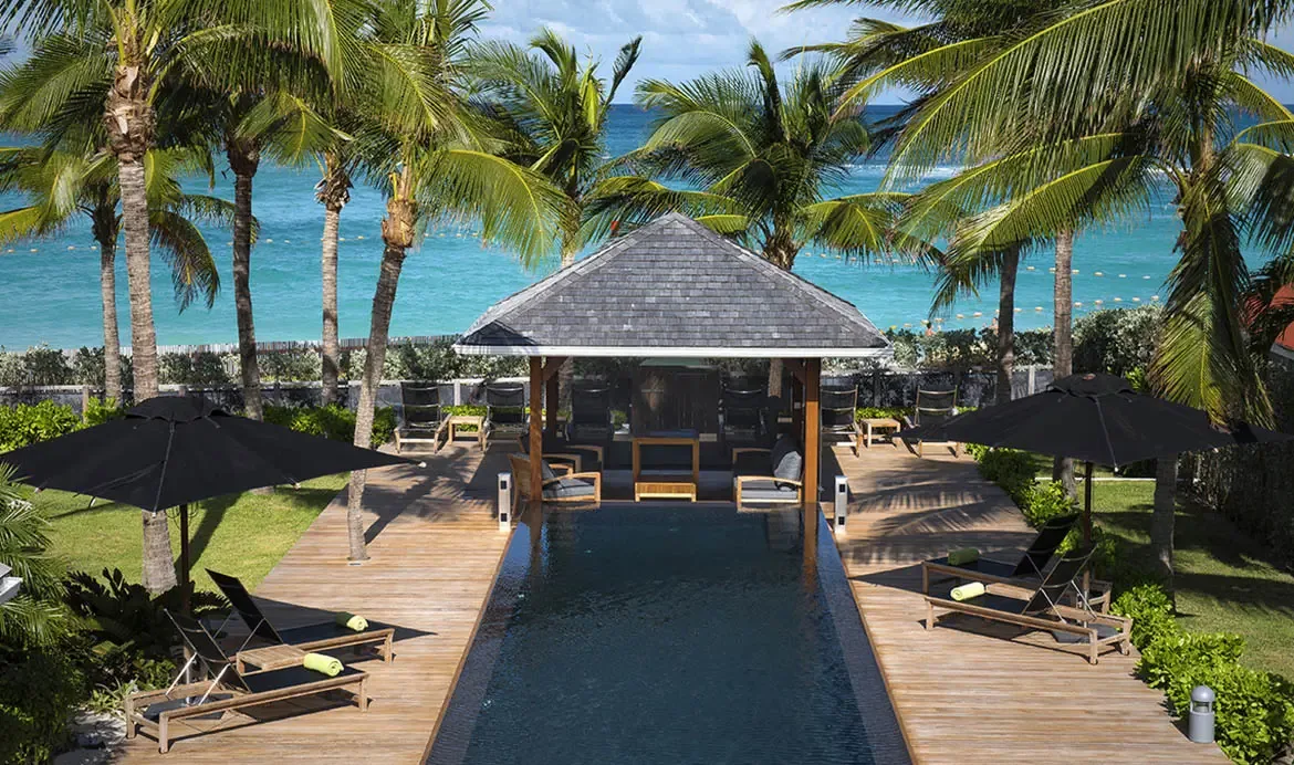 Lime-green towels on sleek loungers line a dark-tiled pool, where a shingled gazebo offers shade and turquoise ocean stretches to the horizon under a bright sky.