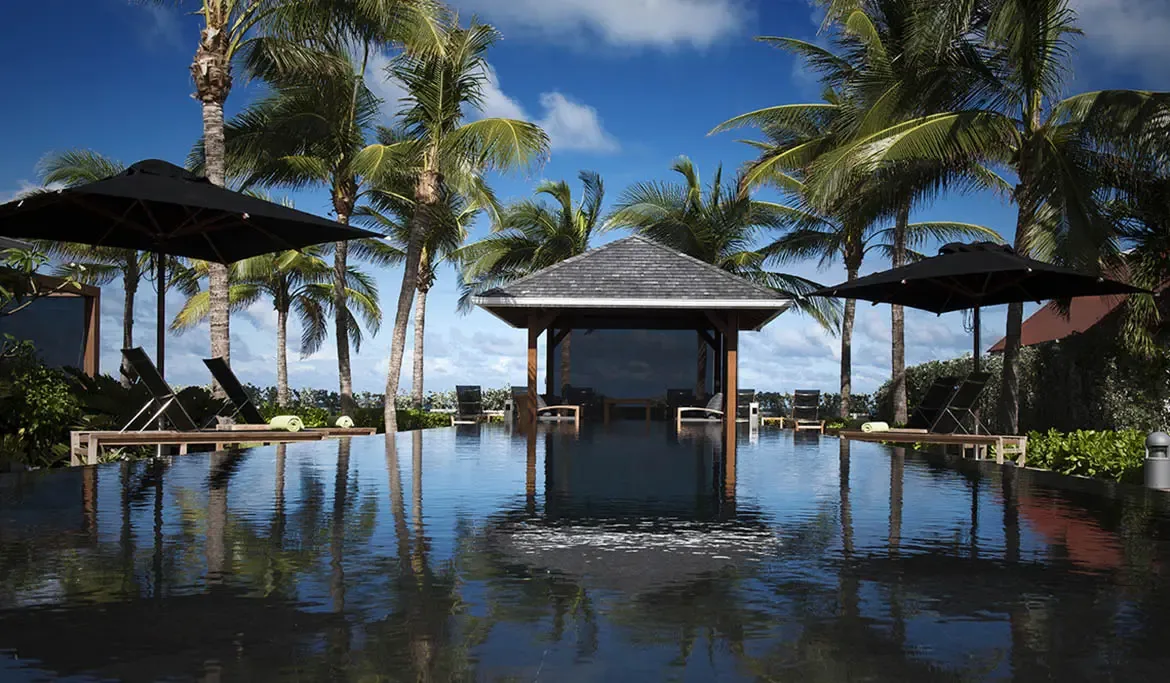 Perfect symmetry: a dark-tiled pool reflects a central gazebo and black umbrellas, while green-towel loungers and palm trees line both sides under a deep blue sky.