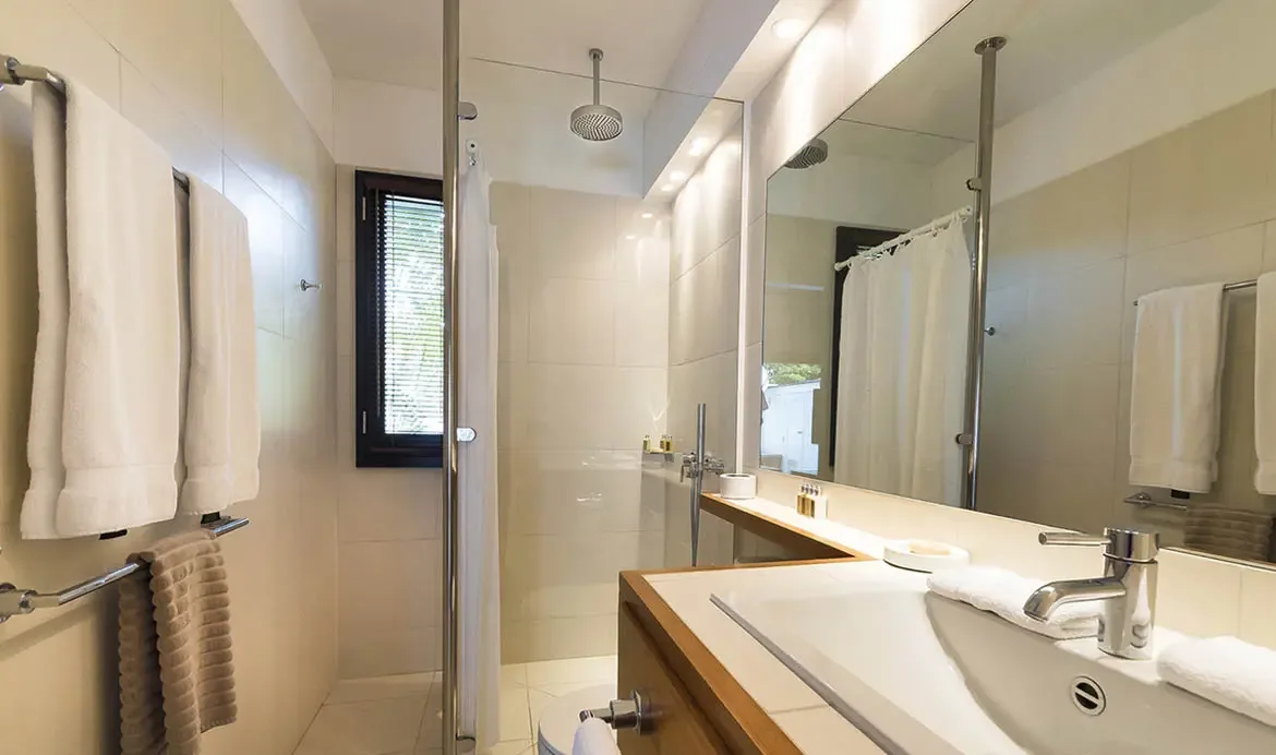 A frameless glass shower with chrome rainfall head pairs with a white wood-edged vanity and a wall-to-wall mirror in this sleek, compact bathroom.