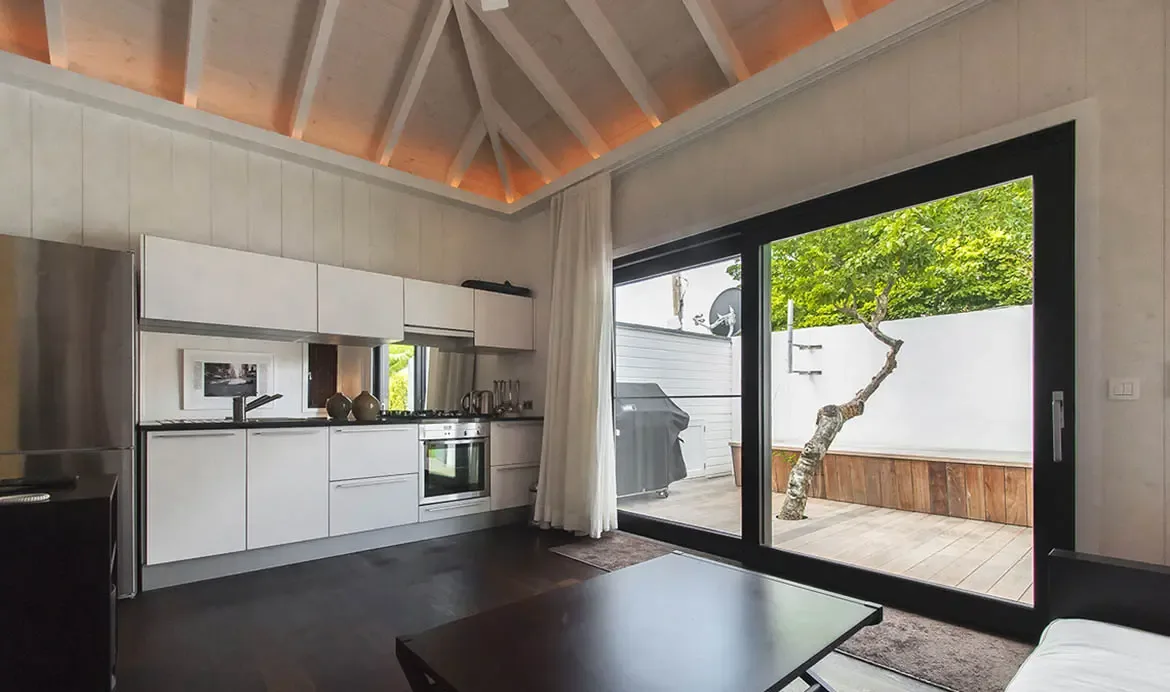 A compact white kitchenette with stainless fridge sits under warm cove lighting, where glass doors open to a wooden deck with a tree and barbecue beyond a white garden wall.