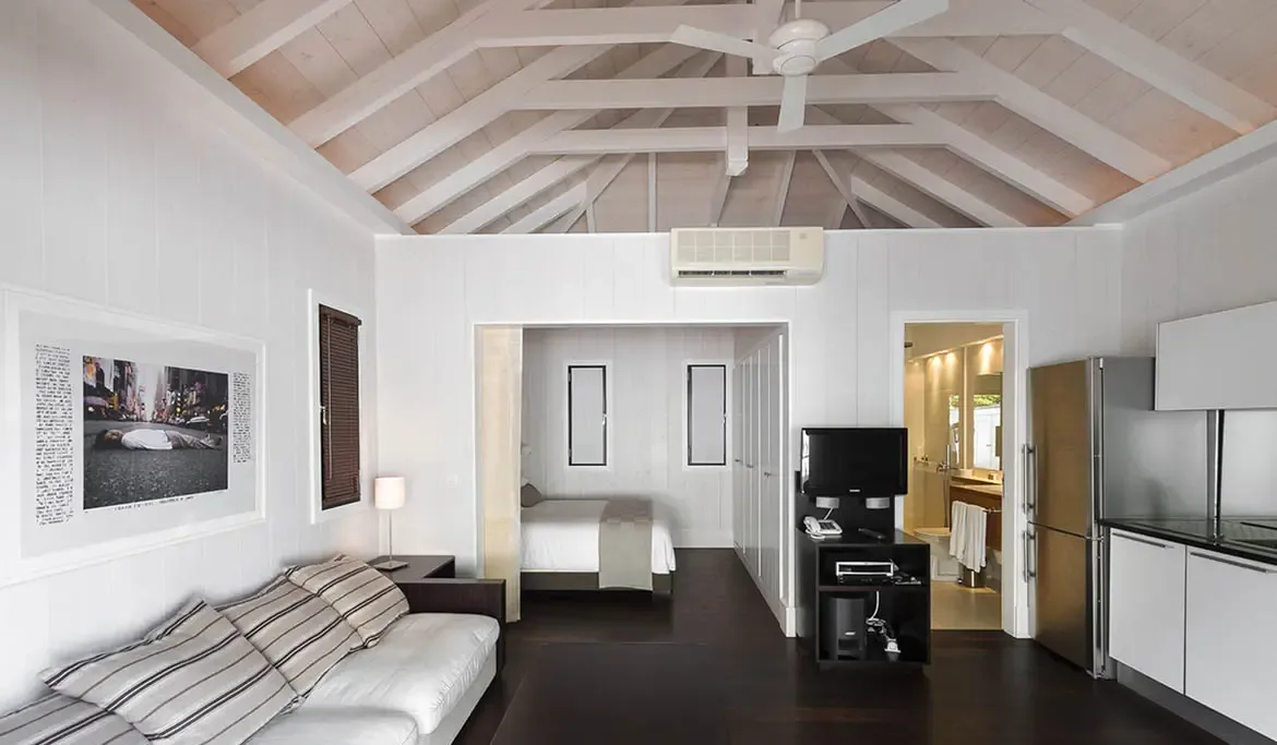 An open-concept studio with a white sofa, kitchenette, and a recessed bed alcove sits under vaulted white rafters, blending living, dining, and sleeping in one bright space.