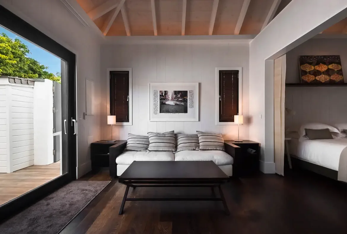 A white sofa with striped pillows faces a vaulted bedroom niche with colorful art, where open doors lead to a private wooden patio enclosed by a white fence and tropical treetops.