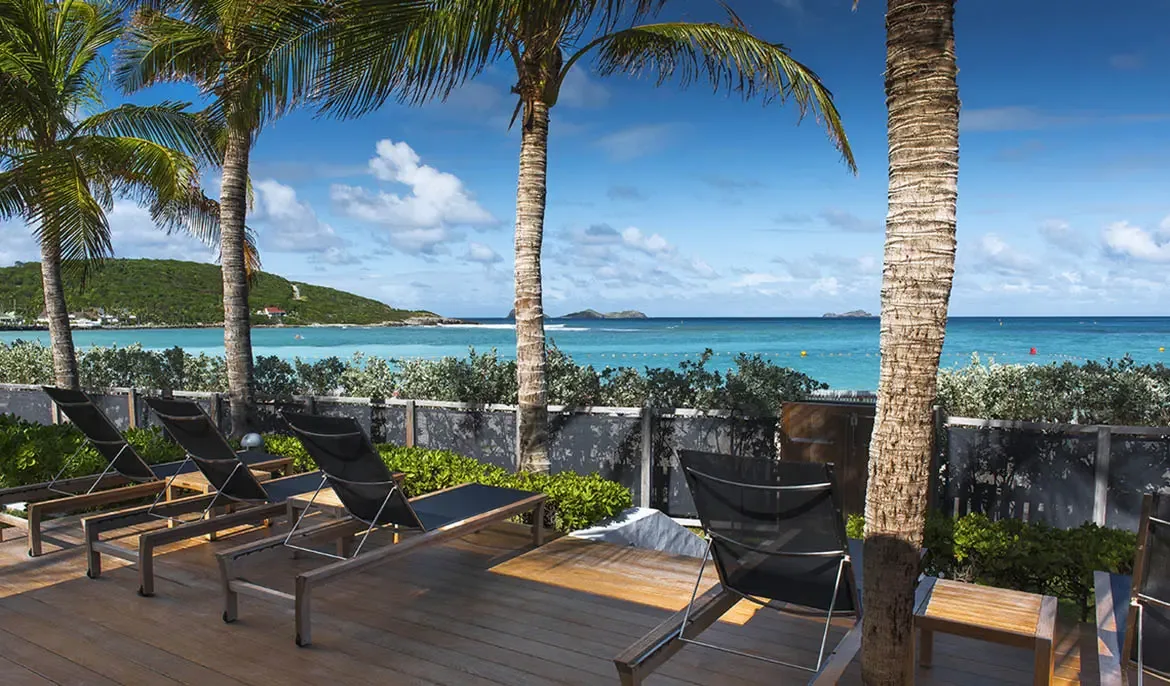 Modern loungers on a wooden deck overlook a calm turquoise bay, where a green hillside dotted with buildings meets the Caribbean and a distant island sits on the horizon.
