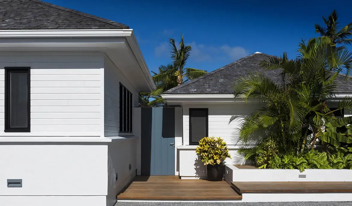 A dusty-blue door offers a subtle accent between white clapboard walls, where tropical greenery fills tiered planters and a wooden deck leads to a bright beachfront entrance.