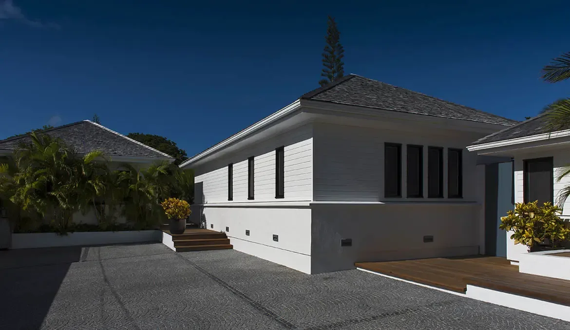White siding and grey-shingled roofs frame a cobblestone courtyard with accent lights, where tropical palms and a tall evergreen rise against a cloudless blue sky.