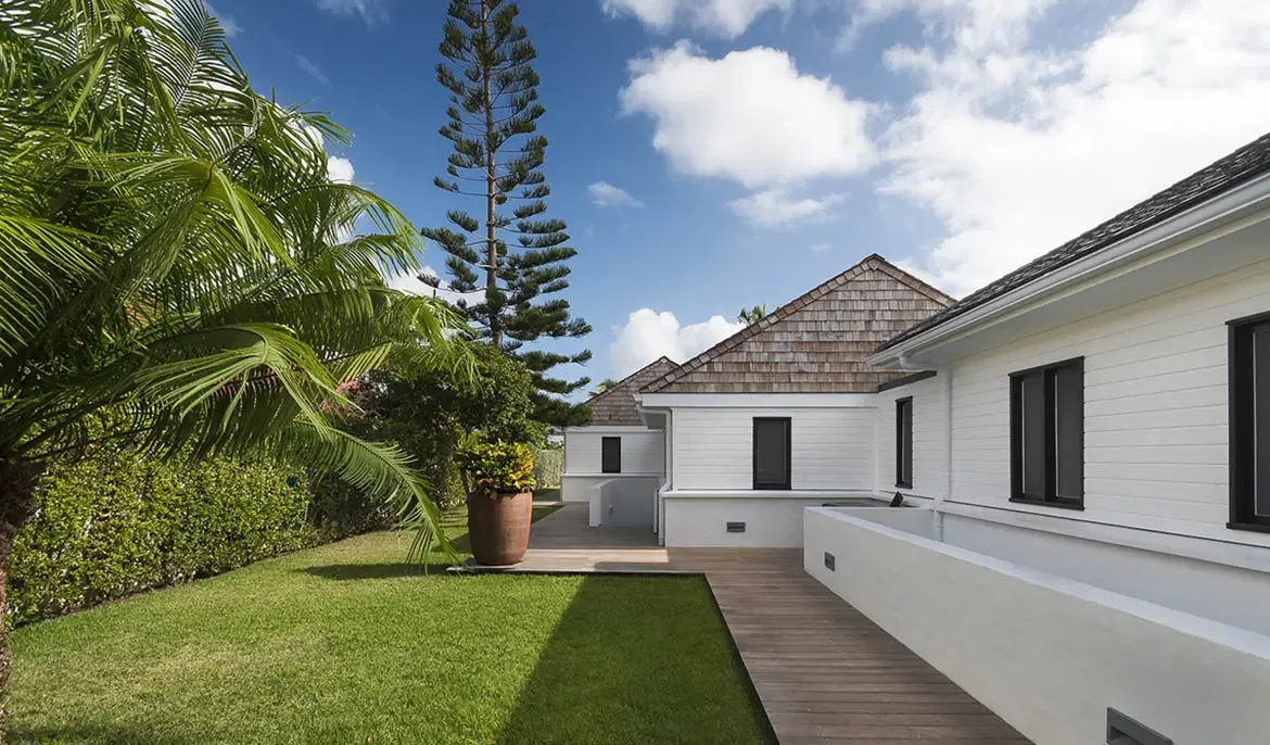 Crisp white siding and dark-framed windows define this modern villa, where a cobblestone courtyard, wooden walkway, and tropical palms create an elegant beachfront arrival.