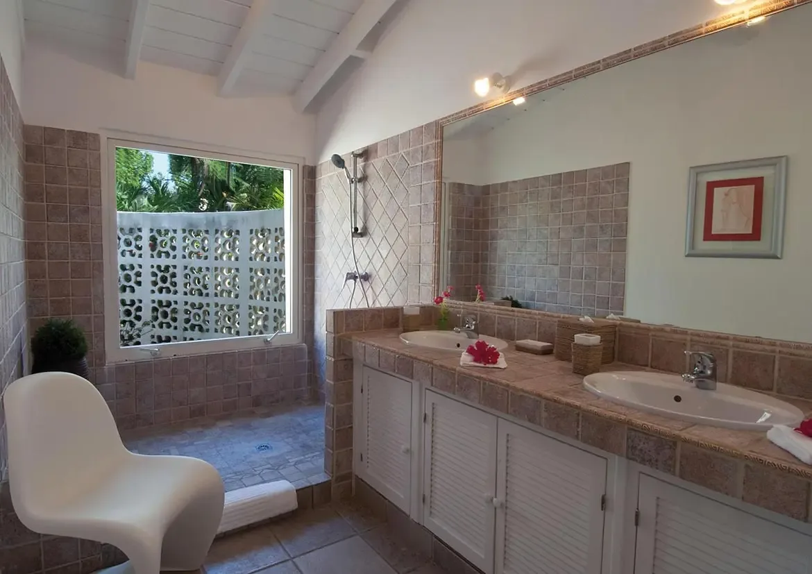 Two white oval sinks with red hibiscus sit on a tan tile double vanity under a frameless mirror, where a walk-in shower and a white modern chair face a garden window.