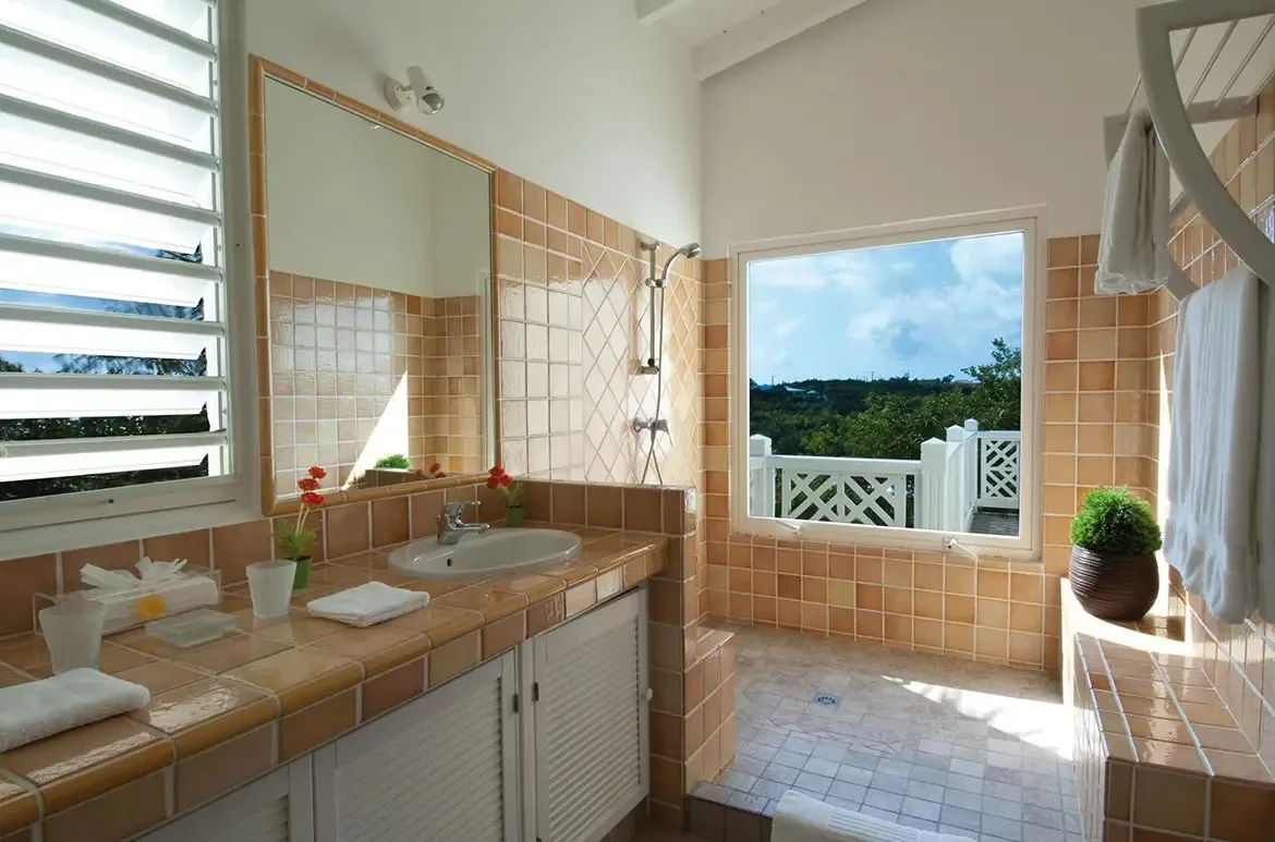 Two white oval sinks with red hibiscus sit on a tan tile vanity beneath a frameless mirror, where a walk-in shower with a garden view opens beside a white modern chair.
