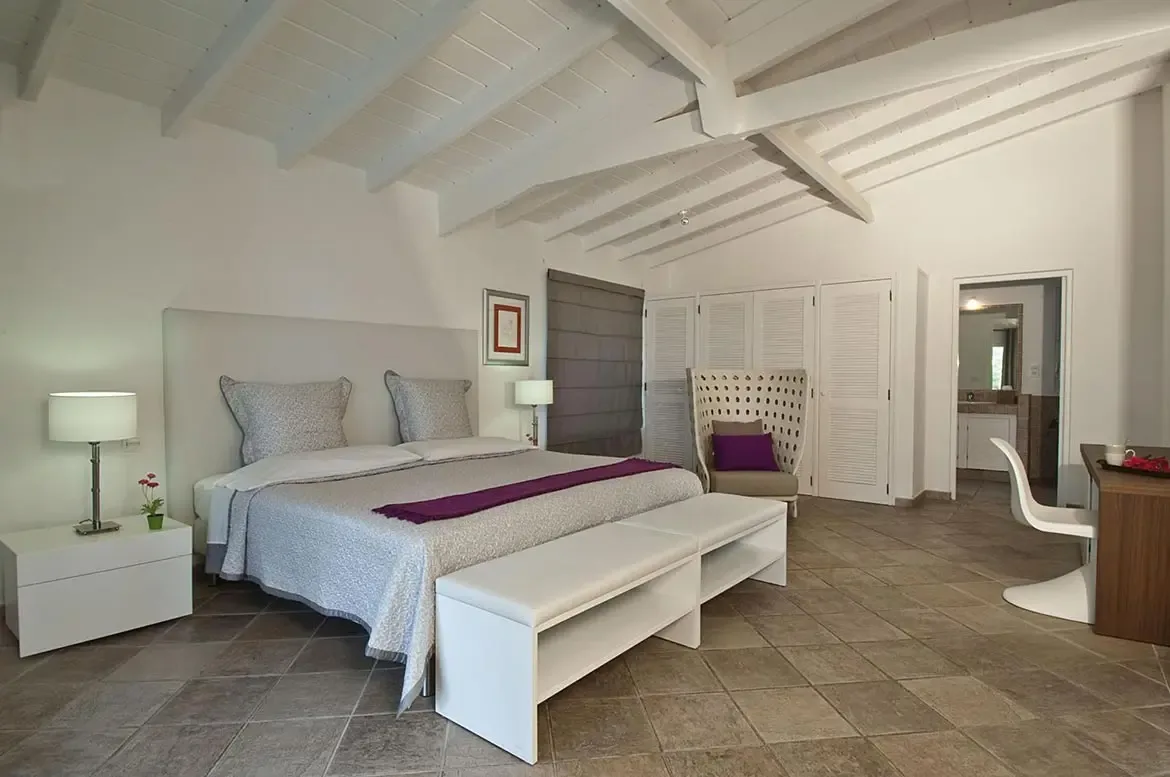 A white headboard bed with a purple runner faces a wicker armchair and louvered closet doors, where a desk with a curved white chair sits beneath a Roman shade.