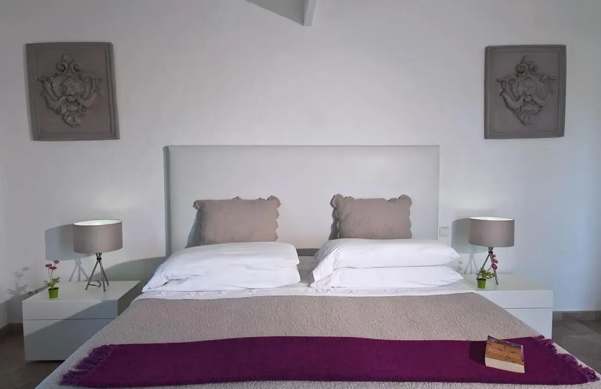 A white headboard bed with a purple throw sits between two matching white nightstands and tripod lamps, where square relief plaques of classical faces flank the headboard above.