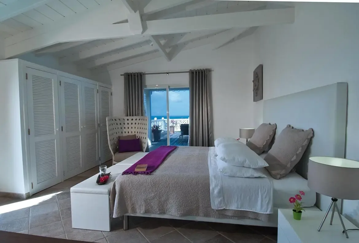 A taupe-quilted bed with a purple throw and pink-flowered tray sits under a vaulted ceiling, where sliding glass doors frame a white-railed terrace and deep blue ocean.