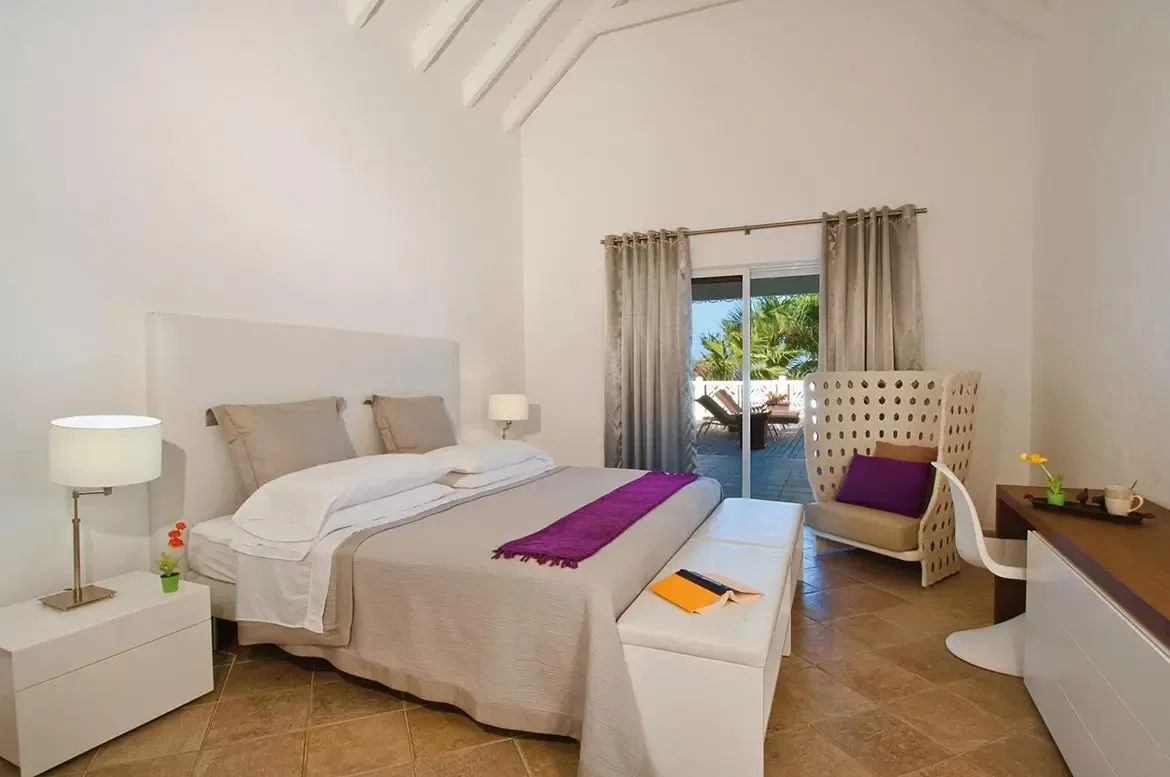 A white headboard bed with a purple runner sits under a vaulted ceiling, where a wicker armchair and open glass doors reveal a terrace with palm trees and ocean.