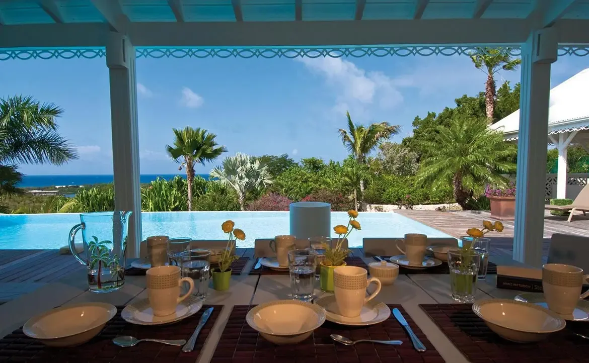 A long table set with white bowls and palm-tree glasses sits under a lattice-trimmed veranda, where a turquoise infinity pool and lush palms overlook a deep blue ocean.