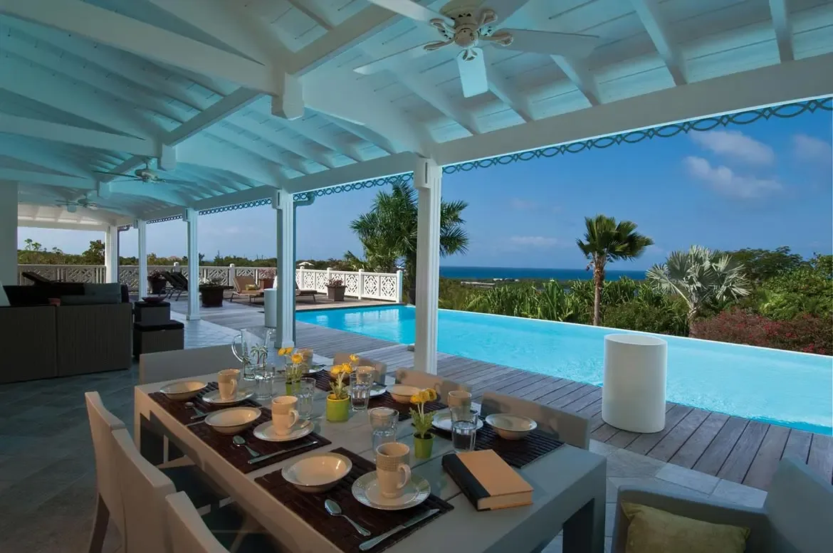 A grey rectangular table with white chairs and yellow flower vases sits under a white pergola, where an infinity pool merges with palms and a deep blue ocean.