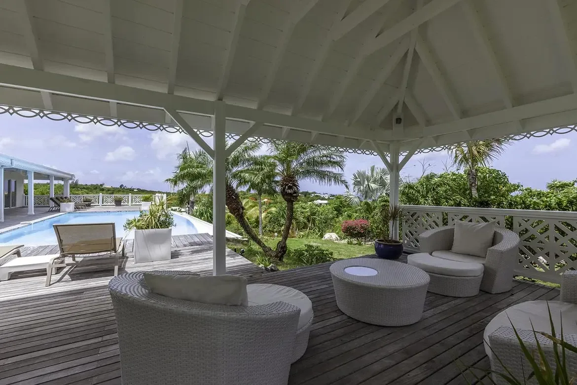 White circular wicker chairs with grey cushions sit under a vaulted white gazebo with scalloped trim, where two tan loungers face a turquoise pool and a deep blue ocean beyond.