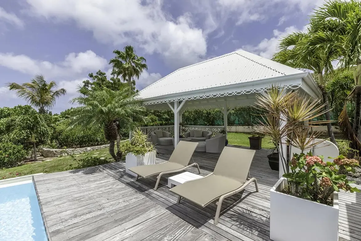 A white gazebo with fretwork trim houses circular wicker chairs and a grey sofa on a manicured lawn, where two beige loungers sit on a weathered deck beside a turquoise pool.