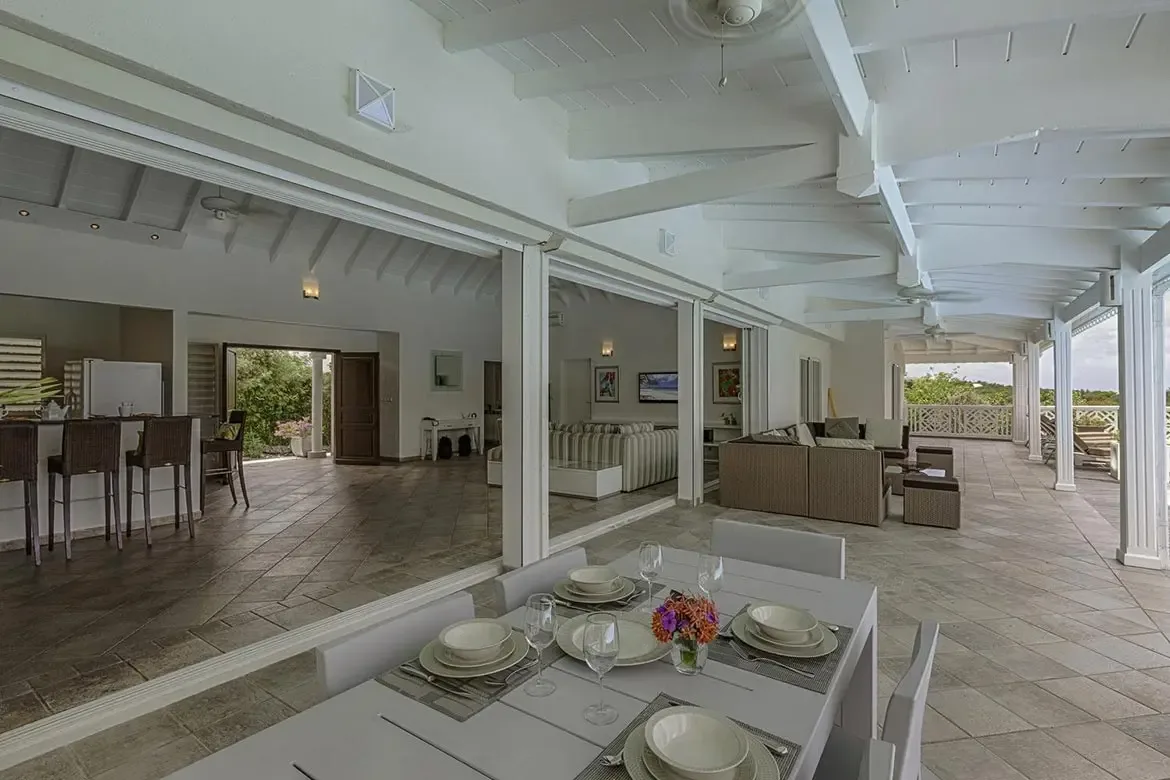 A white dining table set with purple flower arrangements sits under a vaulted ceiling, where open glass doors lead past wicker sofas to a turquoise pool surrounded by tropical greenery.