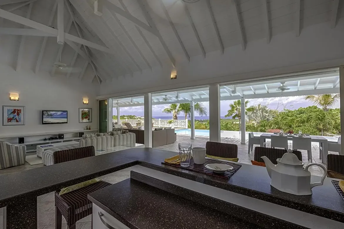 Wicker bar stools sit at a granite counter before a vaulted living area with striped sofas, where open glass doors reveal an outdoor dining table and a turquoise infinity pool.