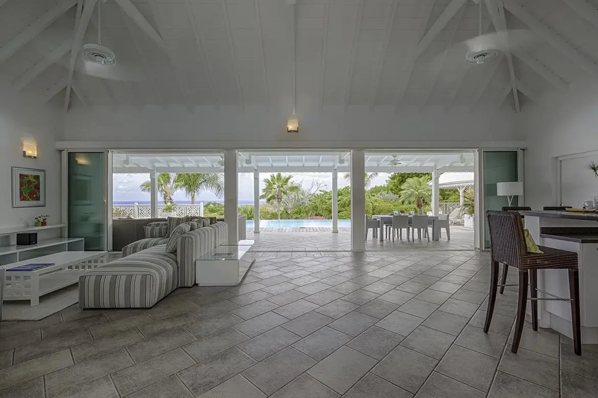 A grey L-shaped sectional sofa faces a flat-screen TV under a vaulted ceiling, where open glass doors reveal a turquoise infinity pool and deep blue ocean beyond lush palms.