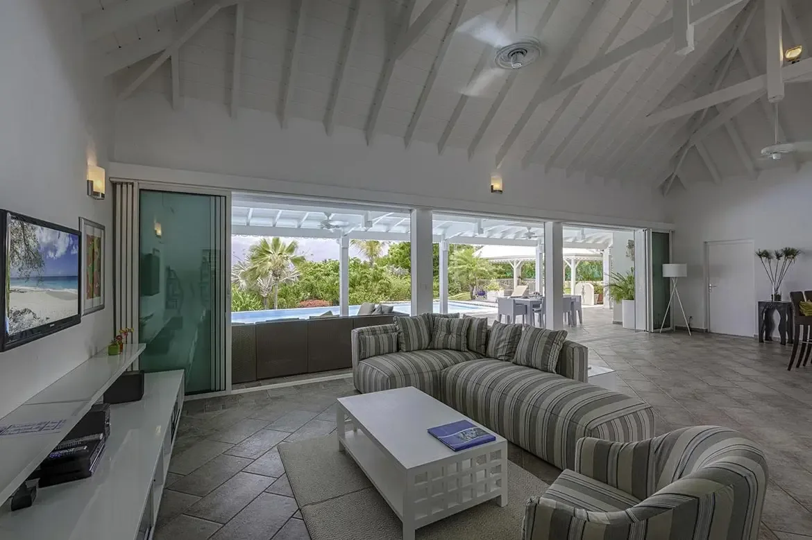 Grey and white striped sofas and an armchair sit on diagonal tiles under a vaulted ceiling, where open glass doors lead to an outdoor dining area and a turquoise pool.