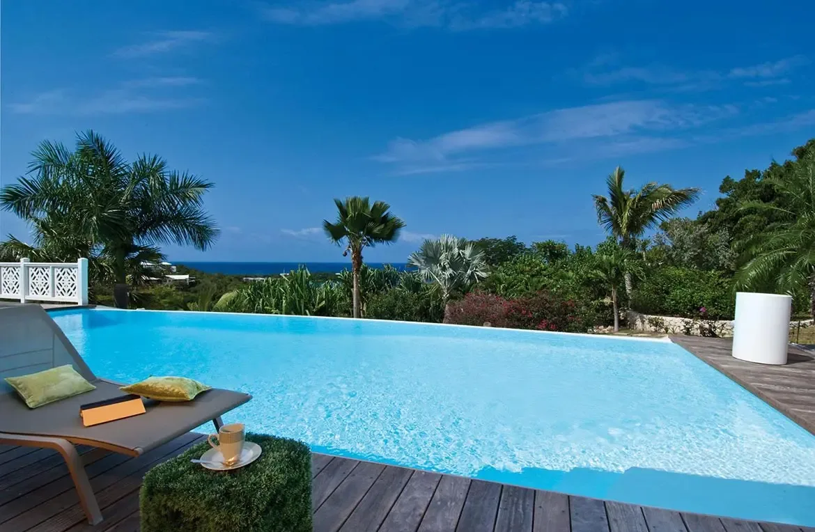 A beige lounger with green pillows and a moss-covered table sits on a dark wood deck beside a turquoise infinity pool, where red bougainvillea and palms frame an ocean view.