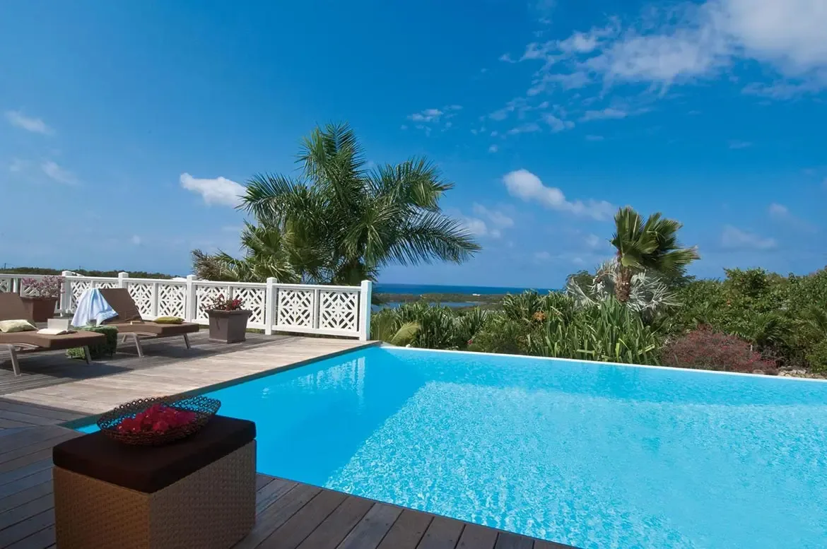 Two wooden loungers with cushions sit on a weathered deck beside a turquoise pool, where a wicker ottoman with magenta bougainvillea and a white lattice railing overlook the ocean.